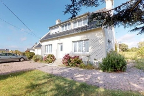 The exterior of a two-story house is shown, featuring a white façade and large windows. A gravel driveway leads to the house, flanked by green shrubs and flowering plants. A vehicle is parked nearby, and a grassy area extends to the side.