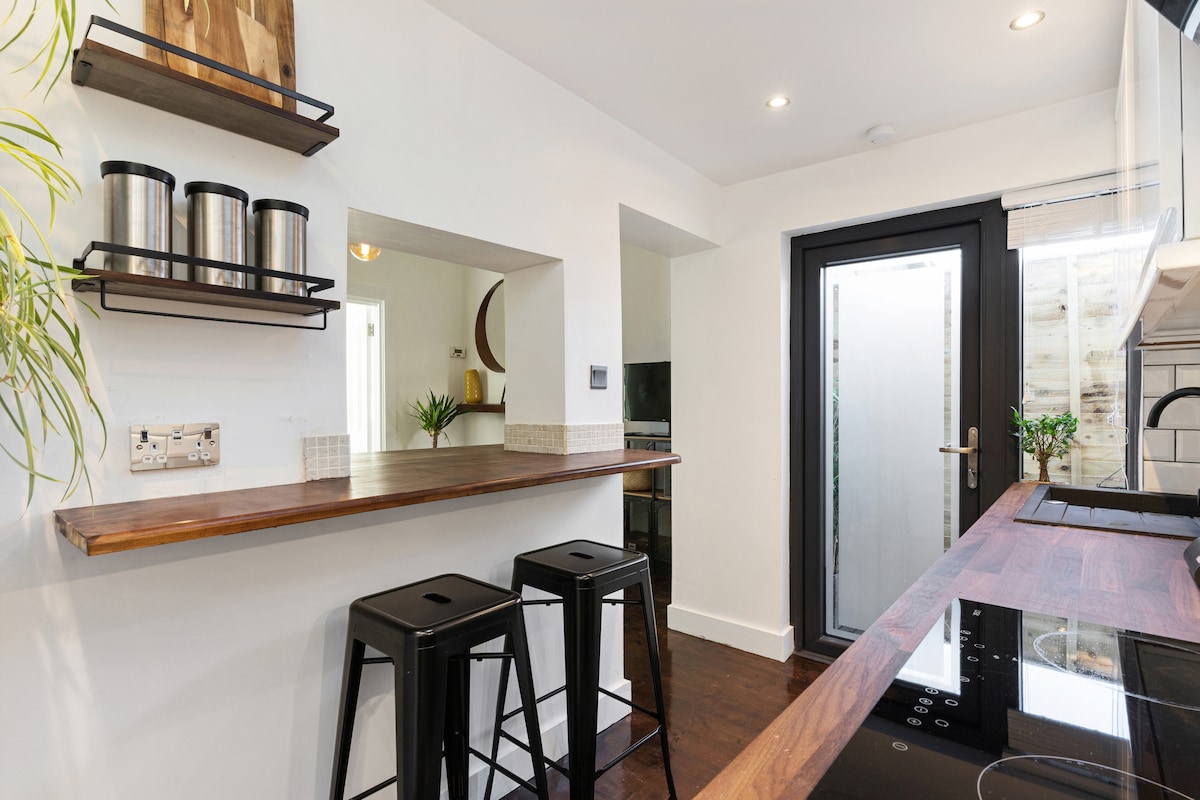 A modern kitchenette is highlighted by sleek countertops and minimalist open shelving. Two black stools are positioned at the bar area, while a glass door allows natural light to enter the space. Plants add a touch of greenery, enhancing the inviting atmosphere.