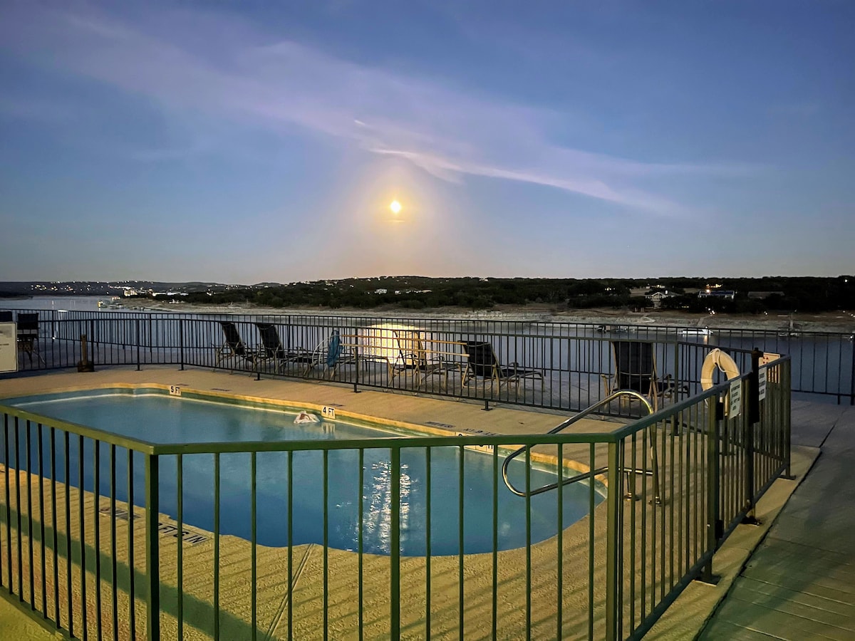 A serene pool area is featured with a view of Lake Travis at dusk. Surrounding the pool, lounge chairs are arranged for relaxation. The moonlight reflects off the water, enhancing the calm atmosphere of the waterfront setting.