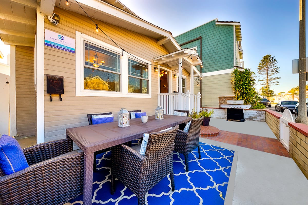 An outdoor dining area is displayed, featuring a rectangular table surrounded by wicker chairs. A blue and white patterned rug adds a decorative touch beneath the furniture. String lights are positioned above, illuminating the space, which is framed by a well-maintained exterior of the house.