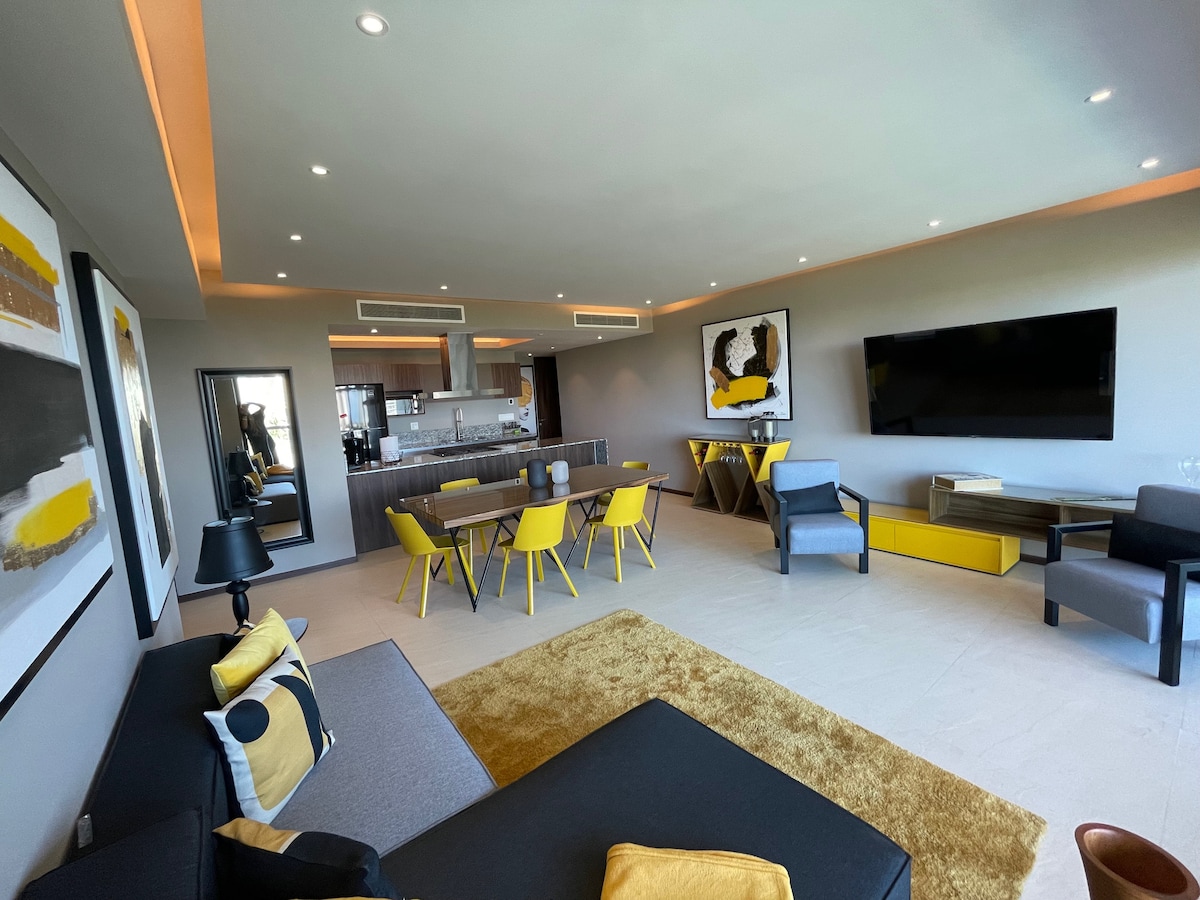 A modern living area features a mix of seating options, including a sectional sofa and dining chairs. Light yellow accents are present throughout, with artworks on the walls. A flat-screen TV is mounted, and the open-concept layout reveals a streamlined kitchen in the background.