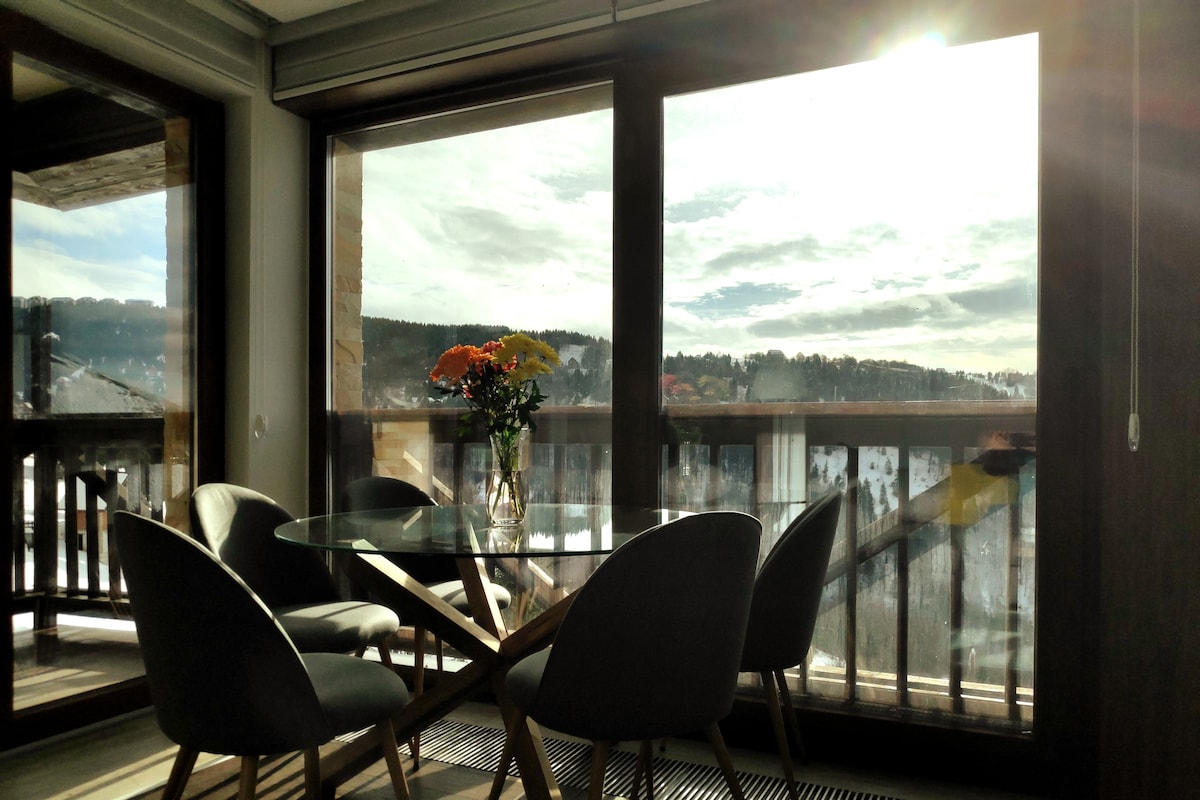 A dining area features a glass table surrounded by four modern chairs. A vase with colorful flowers adds a touch of nature. Large glass doors provide natural light and offer views of the surrounding mountains and landscape.