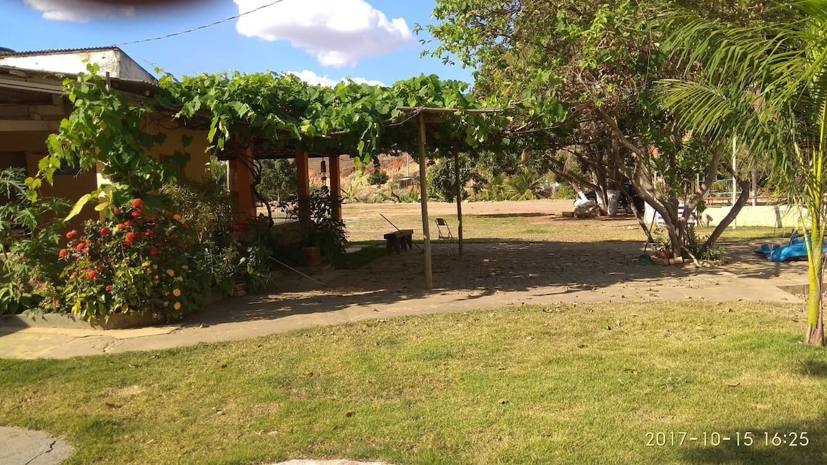 A serene outdoor area is presented, featuring a shaded seating space under a trellis adorned with lush greenery. Colorful flowers bloom nearby, contributing to the peaceful environment. An expanse of grass stretches out, inviting guests to relax and enjoy the natural surroundings.