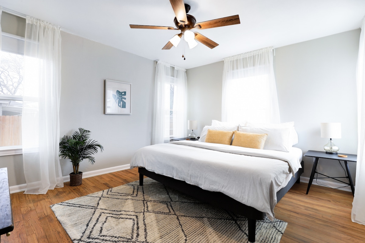 A spacious bedroom features a comfortable bed dressed in crisp linens, flanked by two bedside tables with lamps. Natural light enters through two large windows, while sheer curtains gently frame the views. A patterned area rug lies beneath the bed, enhancing the room's warmth.