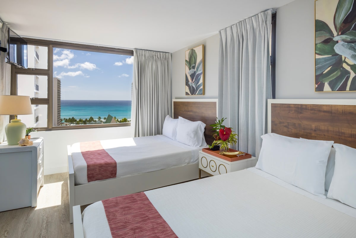 The bright bedroom features two queen beds dressed in crisp white linens, with scenic ocean views visible through large windows. Light-colored curtains frame the windows, and botanical artwork accents the walls. A nightstand with decorative elements and a lamp provides functional aesthetics.