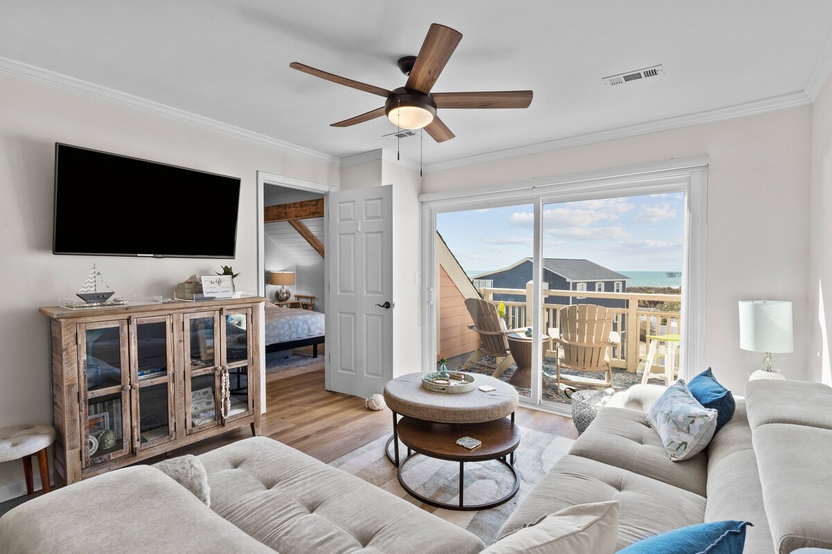 A spacious living area features a soft sectional sofa facing a wall-mounted television. Natural light filters through large sliding glass doors leading to a balcony with outdoor seating. A wooden coffee table rests on a light area rug, complementing the coastal decor.