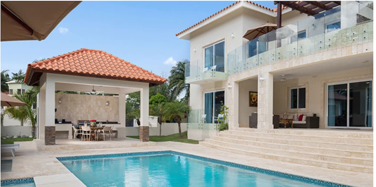 A stunning resort-style villa features a large private pool surrounded by a natural stone deck. The exterior showcases a covered seating area and a spacious terrace, with glass railings offering unobstructed views of the lush garden and inviting outdoor dining area.