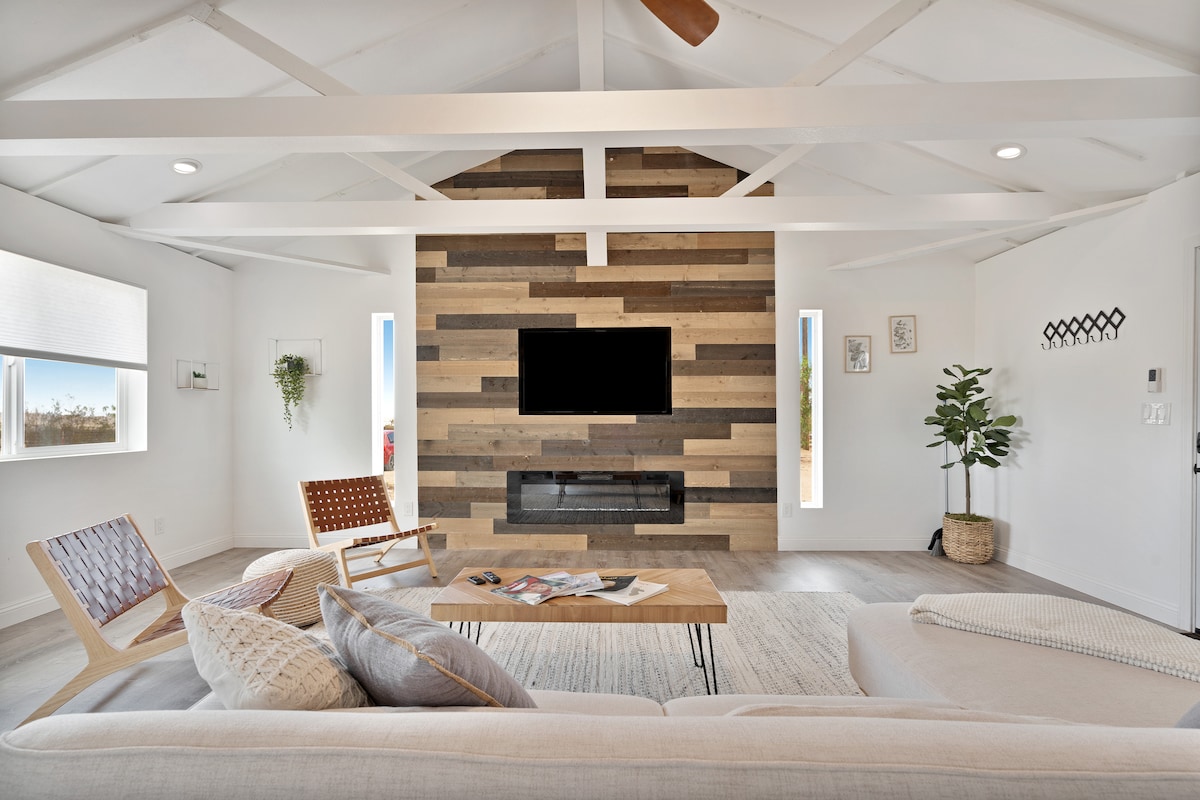 The open-concept living area features a striking barn wood accent wall with a mounted TV. A comfortable sectional sofa and wooden coffee table are arranged for relaxation. Natural light floods through the windows, enhancing the spacious feel of the room.
