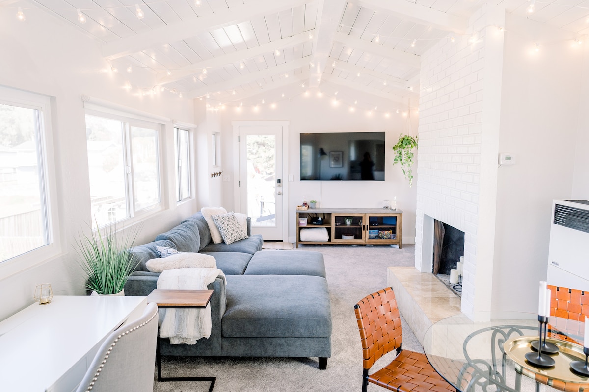 A bright living area features a comfortable sectional sofa under a vaulted ceiling with string lights. Large windows allow natural light, while a television and decorative plants add to the decor. A small dining table and a desk are situated in the space.
