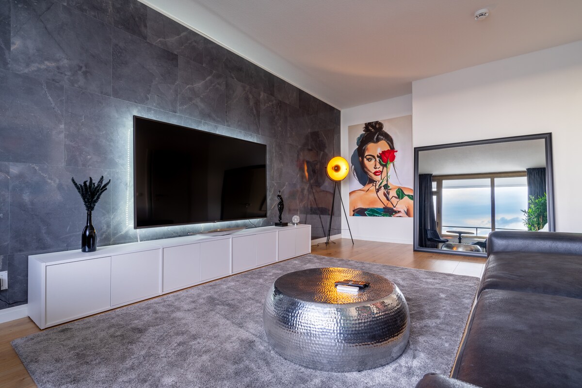 A modern living area features a large flat-screen TV mounted on a textured wall. A sleek coffee table with a metallic finish sits on a plush rug, complemented by a stylish lamp and decorative elements, creating a contemporary yet inviting space.