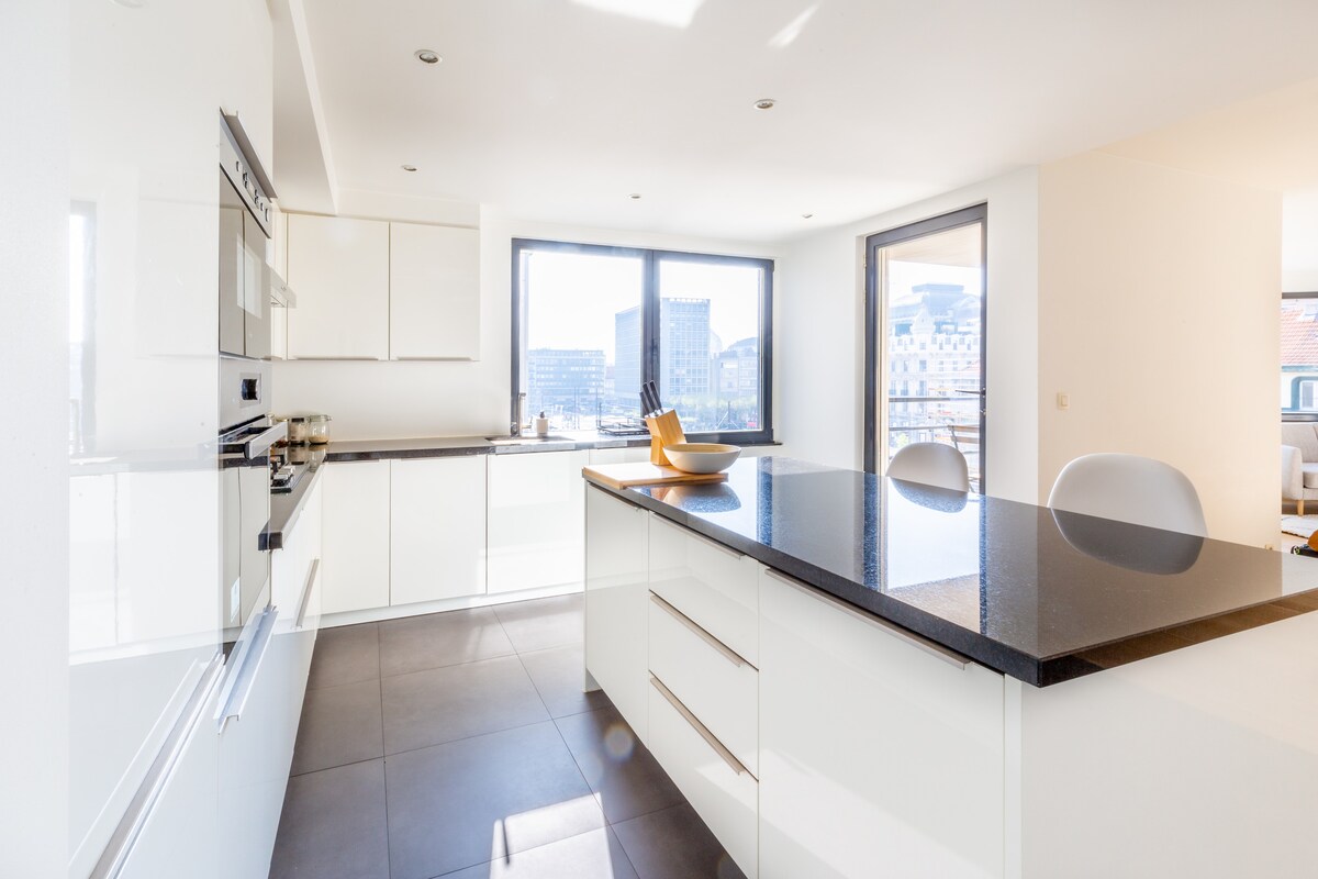 A modern kitchen space features a central island with a dark countertop and light cabinetry. Natural light floods the room through large windows, illuminating sleek appliances and ample counter space. The minimalist design promotes a clean and spacious atmosphere.