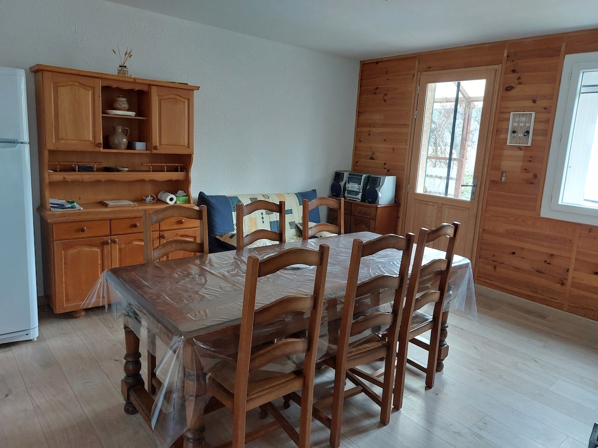A spacious dining area is furnished with a large wooden table surrounded by several chairs. A wooden sideboard is positioned against the wall, displaying various items. A door leads to the outside, while light enters through the window, enhancing the warm wood tones.