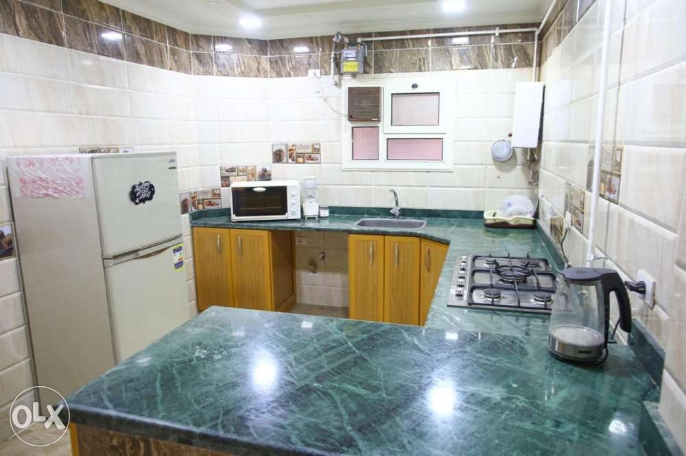 A modern kitchen featuring a green marble countertop with wooden cabinets. Appliances include a refrigerator, microwave, gas stove, and electric kettle. Soft lighting highlights the tiled walls and provides a functional cooking space.