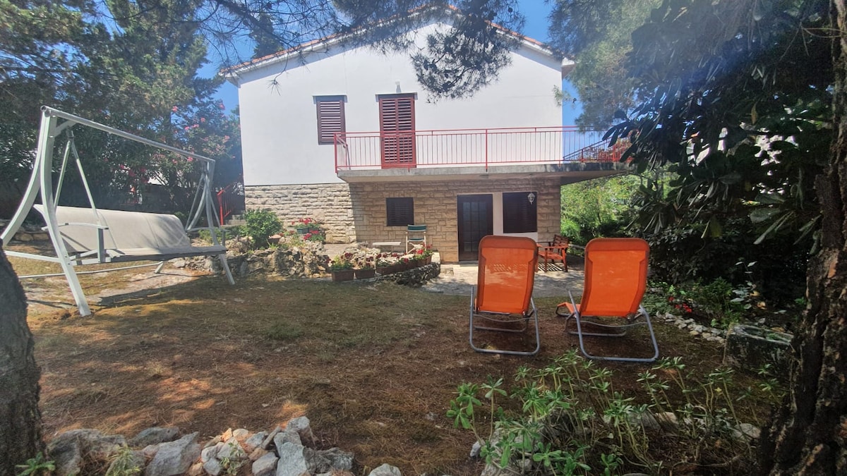 A two-story house features a stone facade and a red balcony overlooking a serene garden. Two orange lounge chairs are arranged on a grassy area, with a swing in the background. Lush greenery and colorful flowers are present, contributing to the peaceful setting.