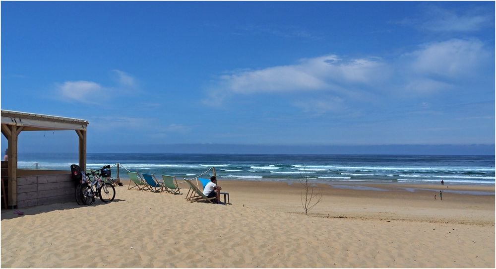 A sandy beach scene extends toward the ocean, with gentle waves lapping at the shore. A few sun loungers are positioned along the beachfront. To the left, bicycles are parked beside a shaded structure, providing a relaxing spot for beachgoers.