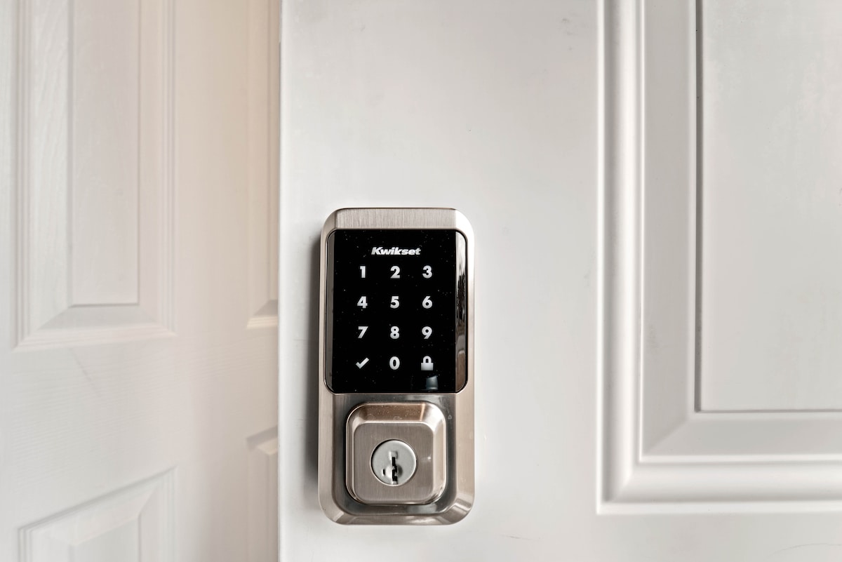 A digital keypad lock is mounted on a white door, showcasing a sleek design. The keypad features numbered buttons and a checkmark, indicating easy access. The door's paneling is detailed, providing a modern touch to the entryway.