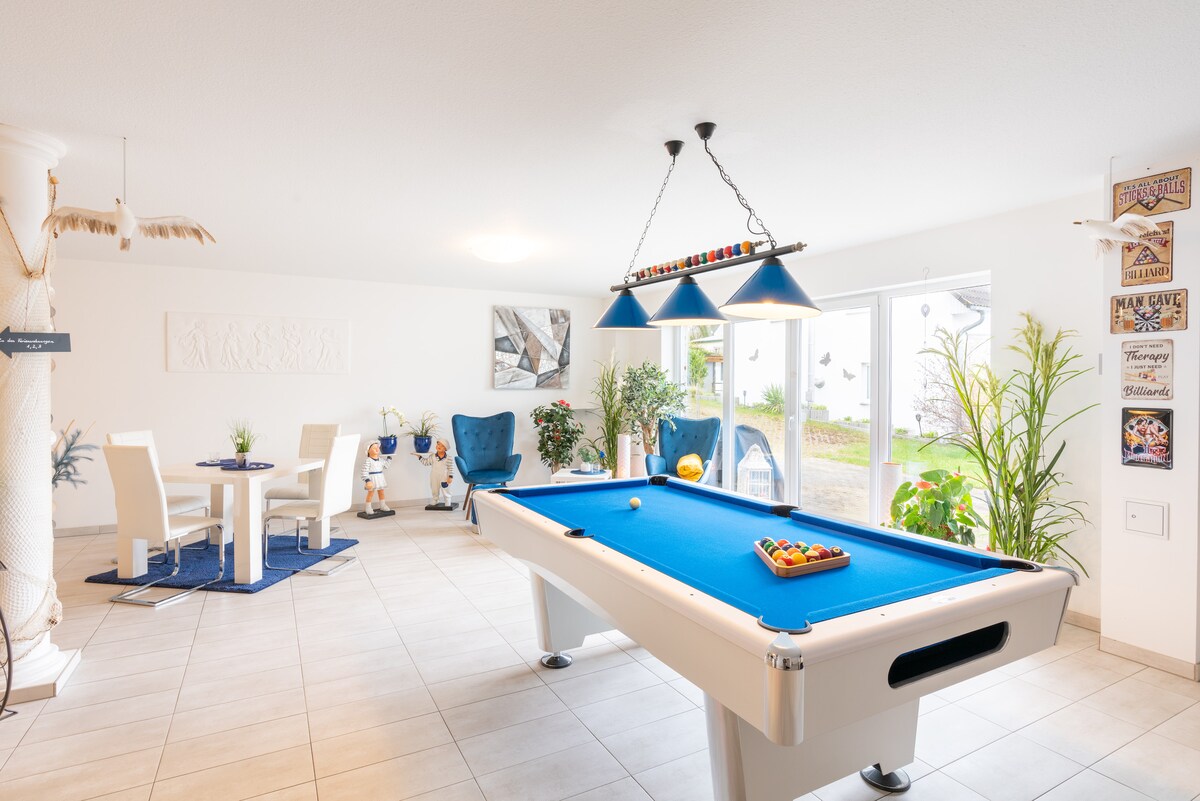 A spacious common area is highlighted by a blue pool table, surrounded by comfortable seating options. Two dining chairs are paired with a round table, while large windows invite natural light and offer views of the outdoors. Decorative plants enhance the inviting decor.