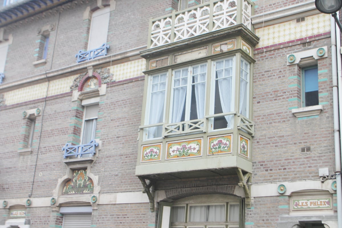 A charming villa exterior is showcased, featuring a distinctive bow window adorned with decorative elements. The building's brick façade is complemented by colorful trim and ornate detailing, including floral motifs. Light drapes gently hang in the window, adding a touch of elegance.