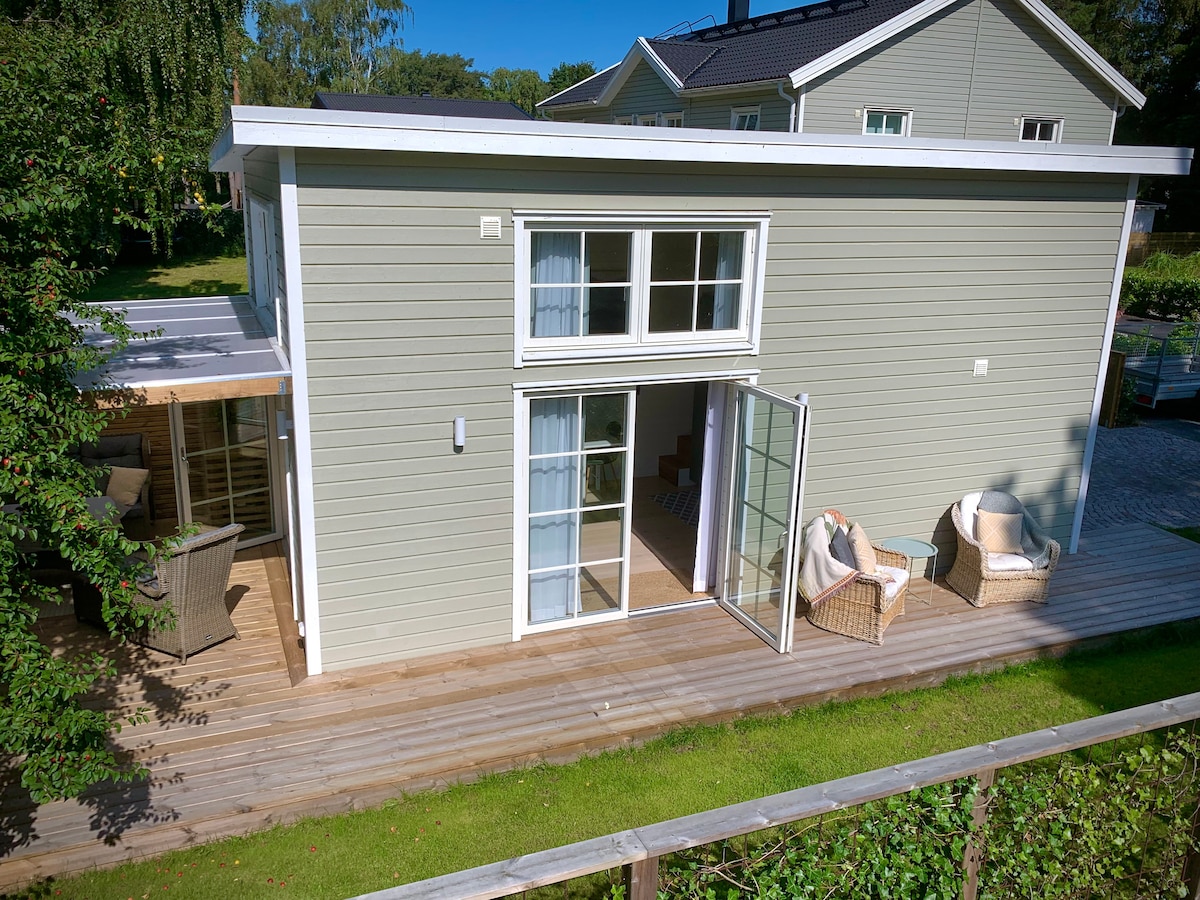 A modern cabin exterior is visible, featuring light green siding and large glass doors that open to a wooden deck. Cozy seating areas are present outside, surrounded by lush greenery, creating a peaceful outdoor space.