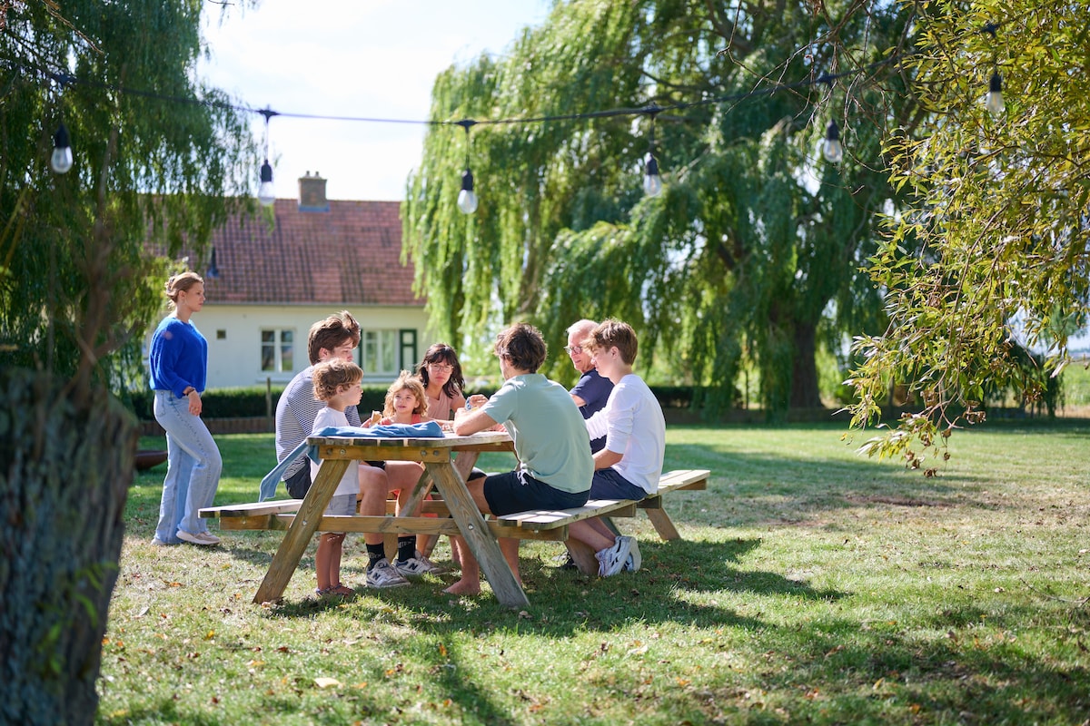 A group of eight individuals gathers around a wooden picnic table in a grassy area. Soft sunlight filters through the trees, and strings of lights are visible overhead. A charming house is situated in the background, framed by lush greenery.