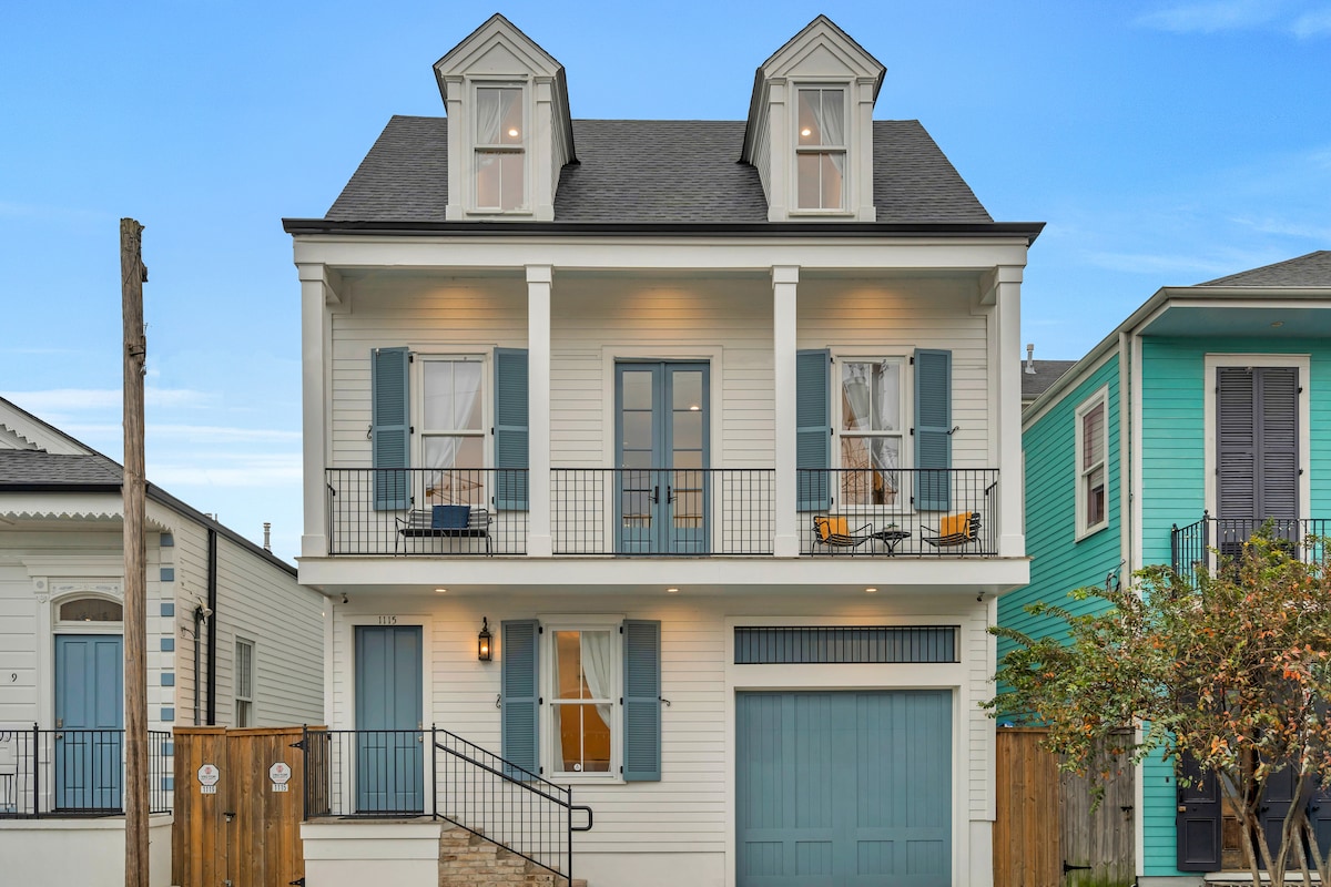 The exterior of a two-story home is showcased, featuring a welcoming façade in soft white with blue shutters. A balcony with seating is visible on the upper level, and the garage is positioned on the ground floor. A well-maintained garden is present at the front.