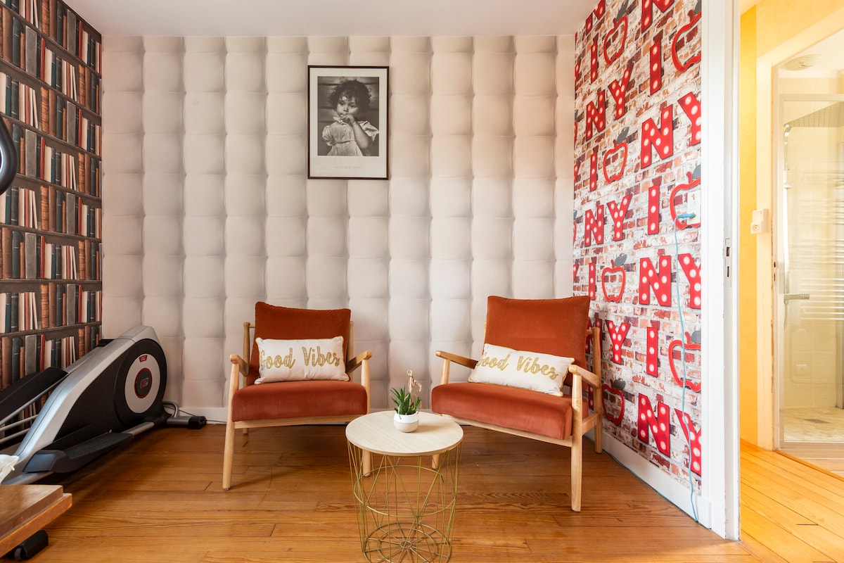 A cozy seating area features two wooden chairs with cushions displaying a positive message, a small round table, and a decorative plant. The textured wall is adorned with a book-patterned wallpaper, while an adjoining wall showcases a vibrant design.