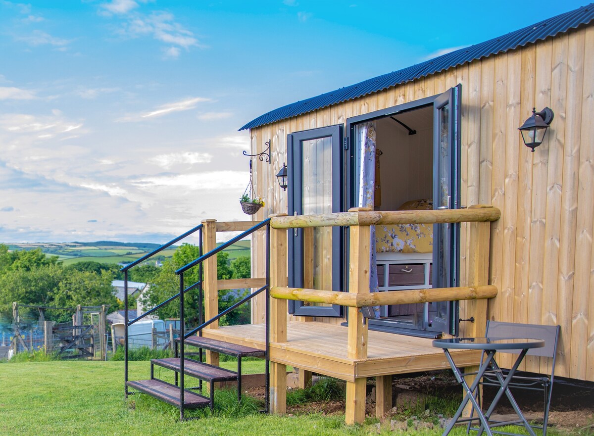 A charming wooden hut is presented with wide double doors opening onto a small deck. A set of stairs leads up to the entrance, and a table and chair are positioned nearby, offering a quiet spot to take in the surrounding views.