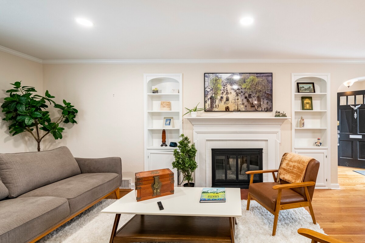 A living room is shown featuring a comfortable grey sofa and a wooden armchair with a light brown seat. A coffee table with a decorative box sits in front of the fireplace, flanked by built-in shelves displaying various décor items. A front door with a window is visible in the background.