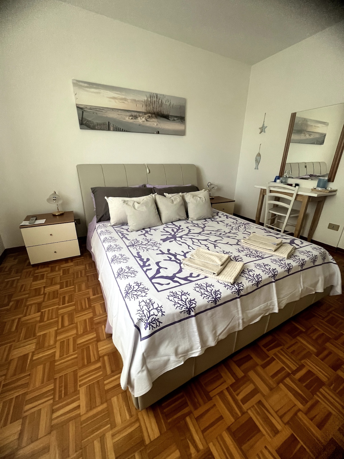 A cozy double room features a spacious bed adorned with decorative pillows and a patterned bedspread. A bedside table and a dresser are present, contributing to a simple and functional layout. Natural light brightens the room, highlighting the warm wooden flooring.