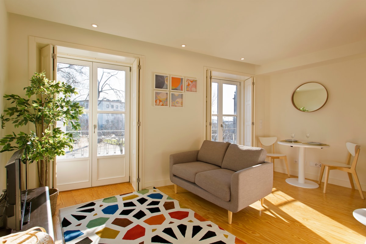 A bright living space features a light-colored sofa arranged near large windows that open to a view of greenery. A colorful geometric rug adds interest to the wooden floor. A round dining table with two chairs is set against the wall, enhancing the room's inviting atmosphere.