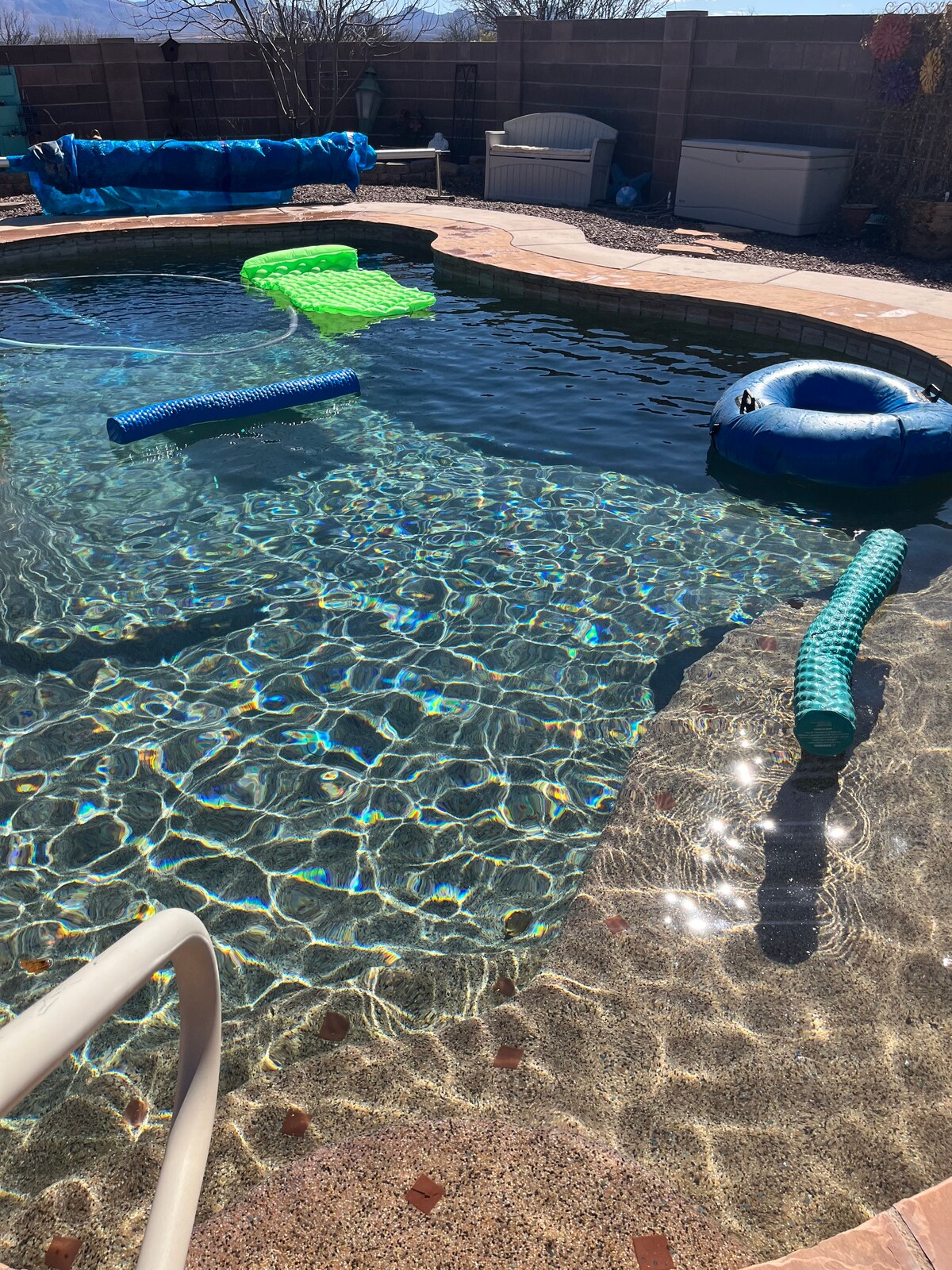 A refreshing pool area is depicted, showcasing clear blue water accented by vibrant green and blue pool floats. Sunlight reflects off the water's surface, creating a shimmering effect. Surrounding the pool are areas of stone decking and a gated barrier in the background.
