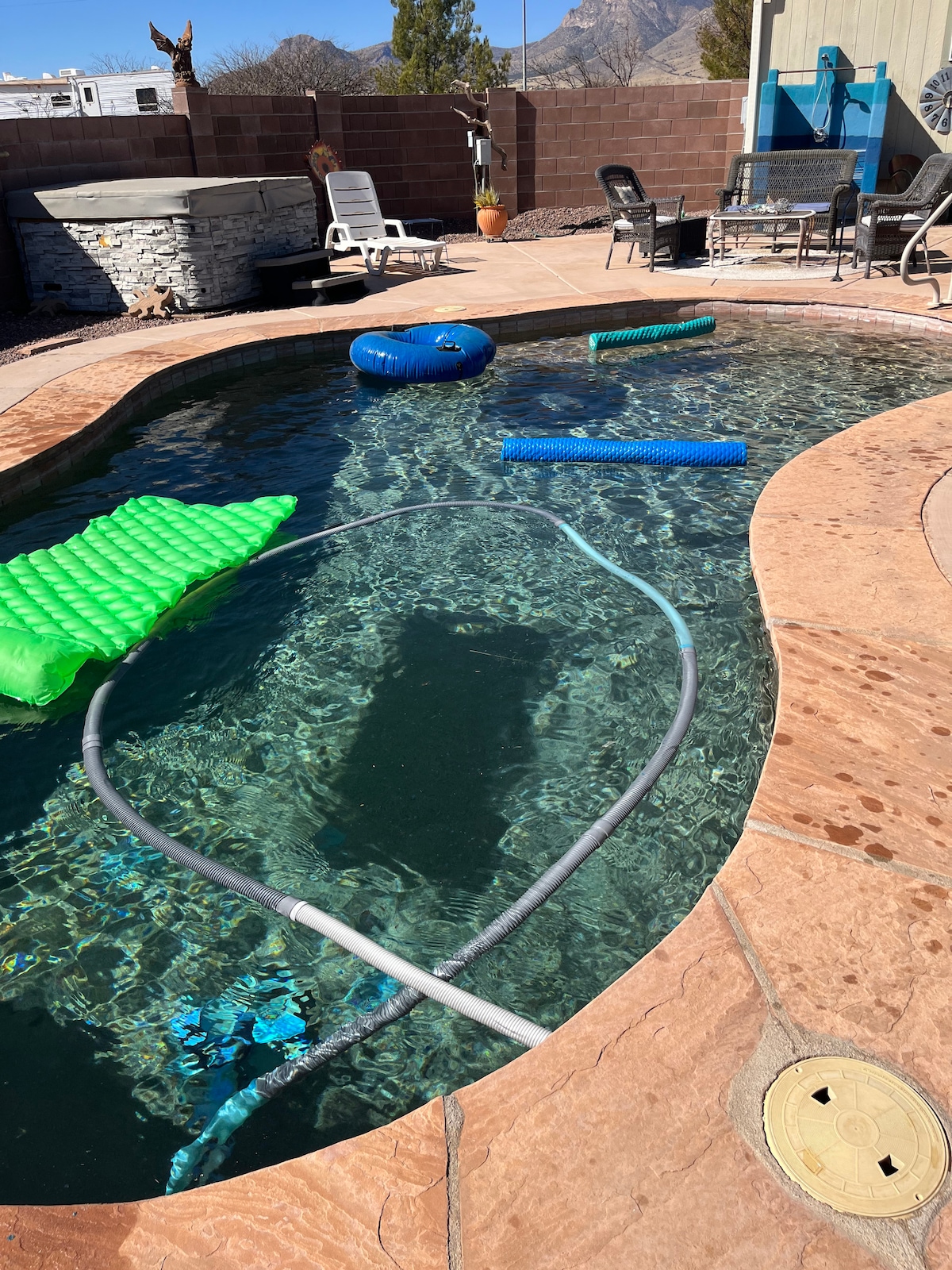 A swimming pool is set within a fenced area, featuring floating loungers and pool toys in vibrant green and blue colors. The pool's surface gleams under sunlight, with a backdrop of rugged mountains visible in the distance.