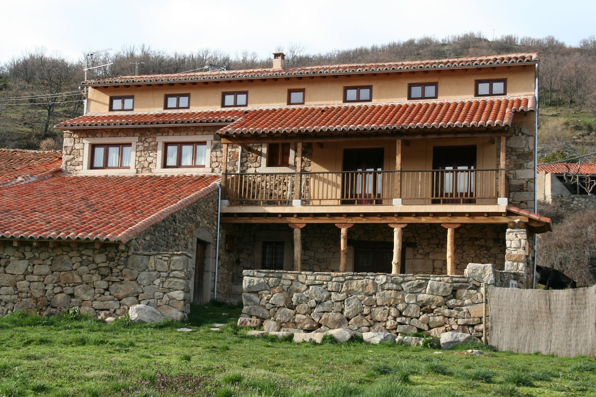 A two-story rustic house is set against a hillside, featuring a combination of stone and wood construction. The terracotta roof is complemented by multiple windows. A balcony extends from the upper level, providing views of the surrounding landscape. The grassy foreground adds to the natural setting.