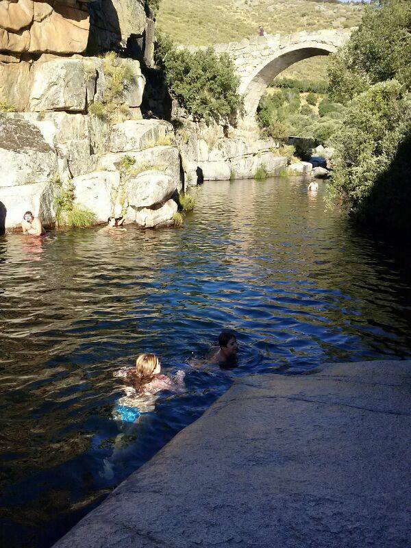 A natural swimming area is surrounded by rocky cliffs and lush greenery. Two individuals are seen swimming in the clear water, while an arched stone bridge is visible in the background, enhancing the serene landscape.