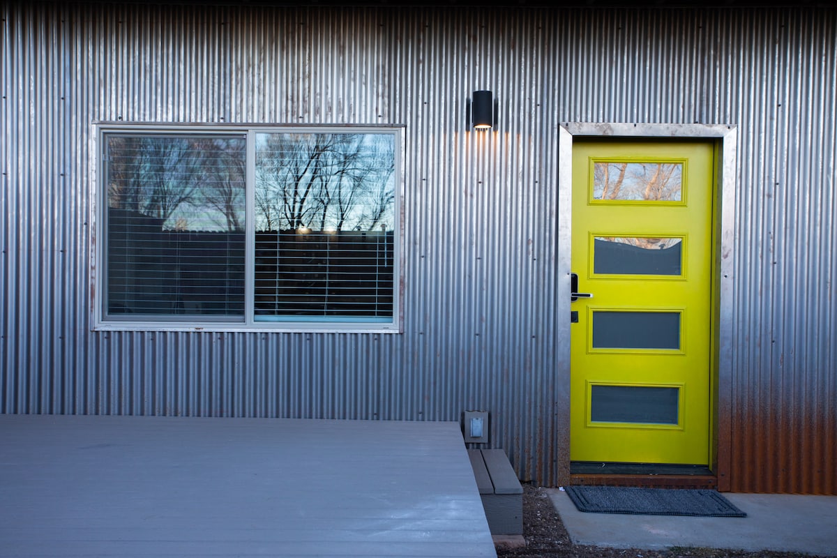 A striking green door complements a corrugated metal exterior. A large window with horizontal blinds reflects the surrounding trees. A small welcoming porch offers a space to pause before entering the unit.