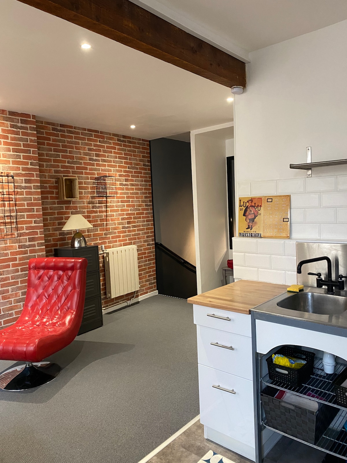 A bright entrance area features a stylish red chair against a backdrop of exposed brick walls. White cabinetry and a stainless-steel sink are visible in the kitchen space, while warm lighting enhances the ambiance. A staircase leads to an upper level, inviting exploration.