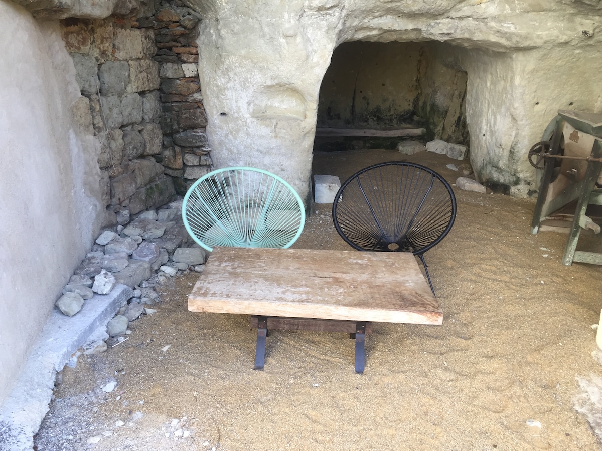 An intimate setting is presented with two modern chairs, one in mint green and the other in black, situated around a rustic wooden table. The natural stone walls of the troglodyte cave create an earthy backdrop, enhancing the unique ambiance of the space.