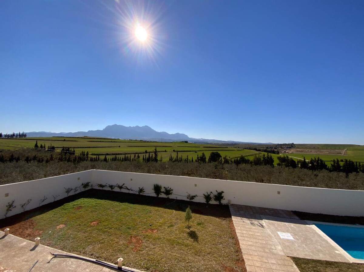 A clear blue sky filled with sunlight overlooks expansive fields and distant mountains. The view captures a serene landscape, framed by green foliage and a neatly maintained garden. A swimming pool is visible adjacent to the terrace, providing an inviting outdoor space.