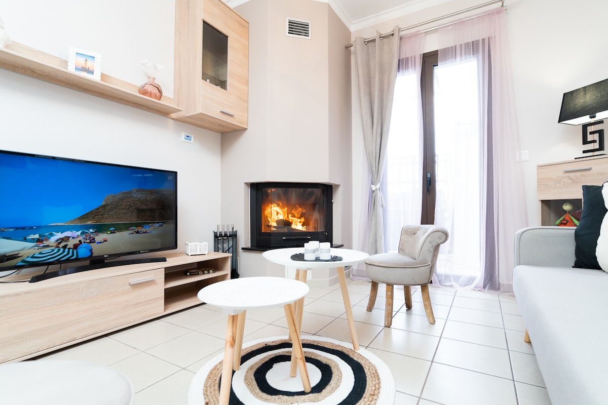 A cozy living area is highlighted by a functional fireplace, accompanied by a flat-screen television. Two small tables are placed on a round area rug, and a comfortable chair is positioned nearby, while natural light filters through the glass doors leading outside.