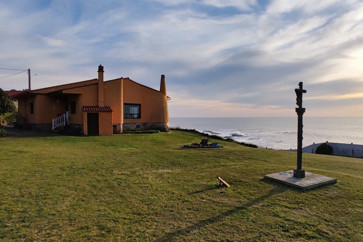 A charming property is situated on a grassy area overlooking the sea, featuring a modest yellow house with a red roof. A stone cross stands prominently on the lawn, while the ocean is visible in the background under a sky filled with soft clouds.