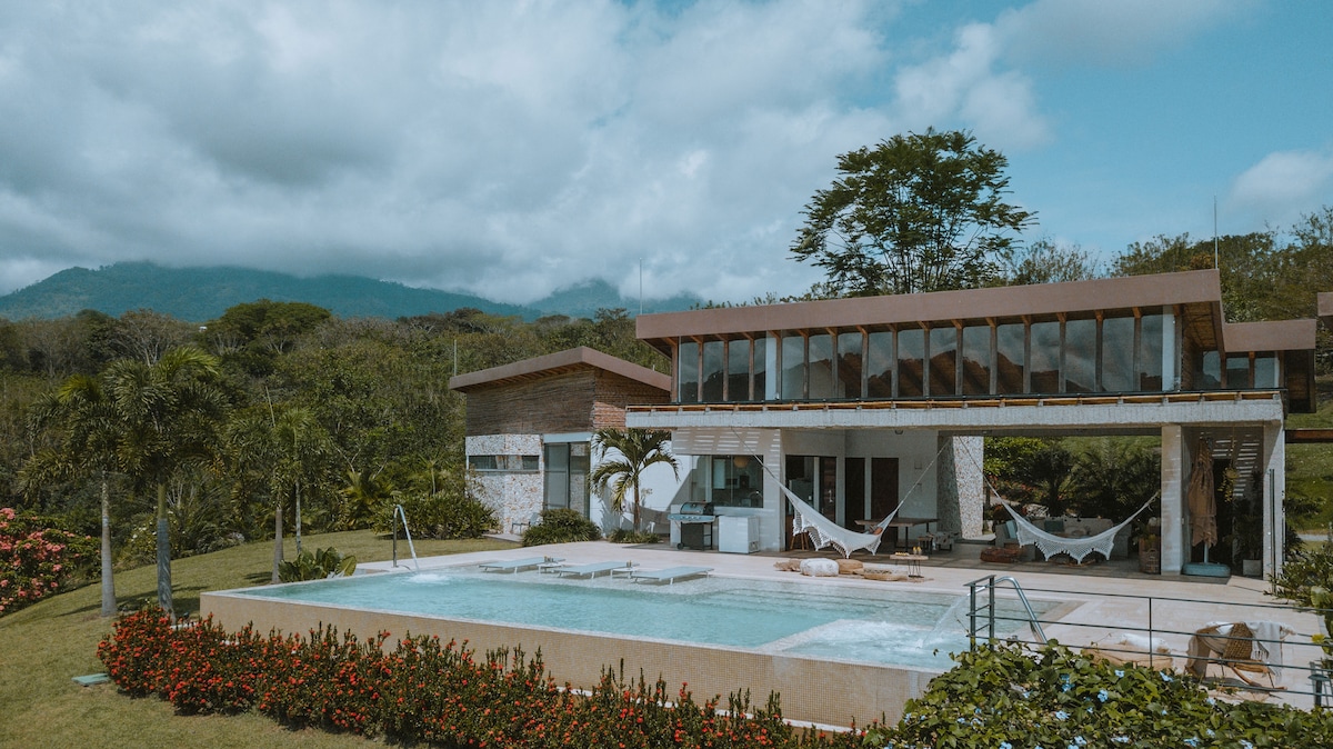 A modern villa is framed by lush greenery, featuring a spacious pool with a jacuzzi nearby. Hammocks are elegantly suspended for relaxation. Large windows invite natural light into the open layout, while mountains provide a serene backdrop.