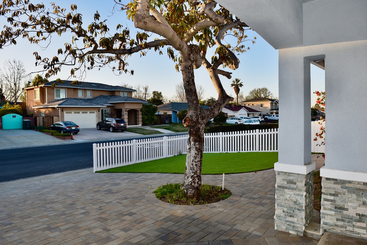 A view of the property's exterior showcases a well-maintained front yard with a manicured lawn and a decorative tree. A white picket fence outlines the property, with a peaceful street visible in the background. Nearby, residential houses and cars are situated along the road.