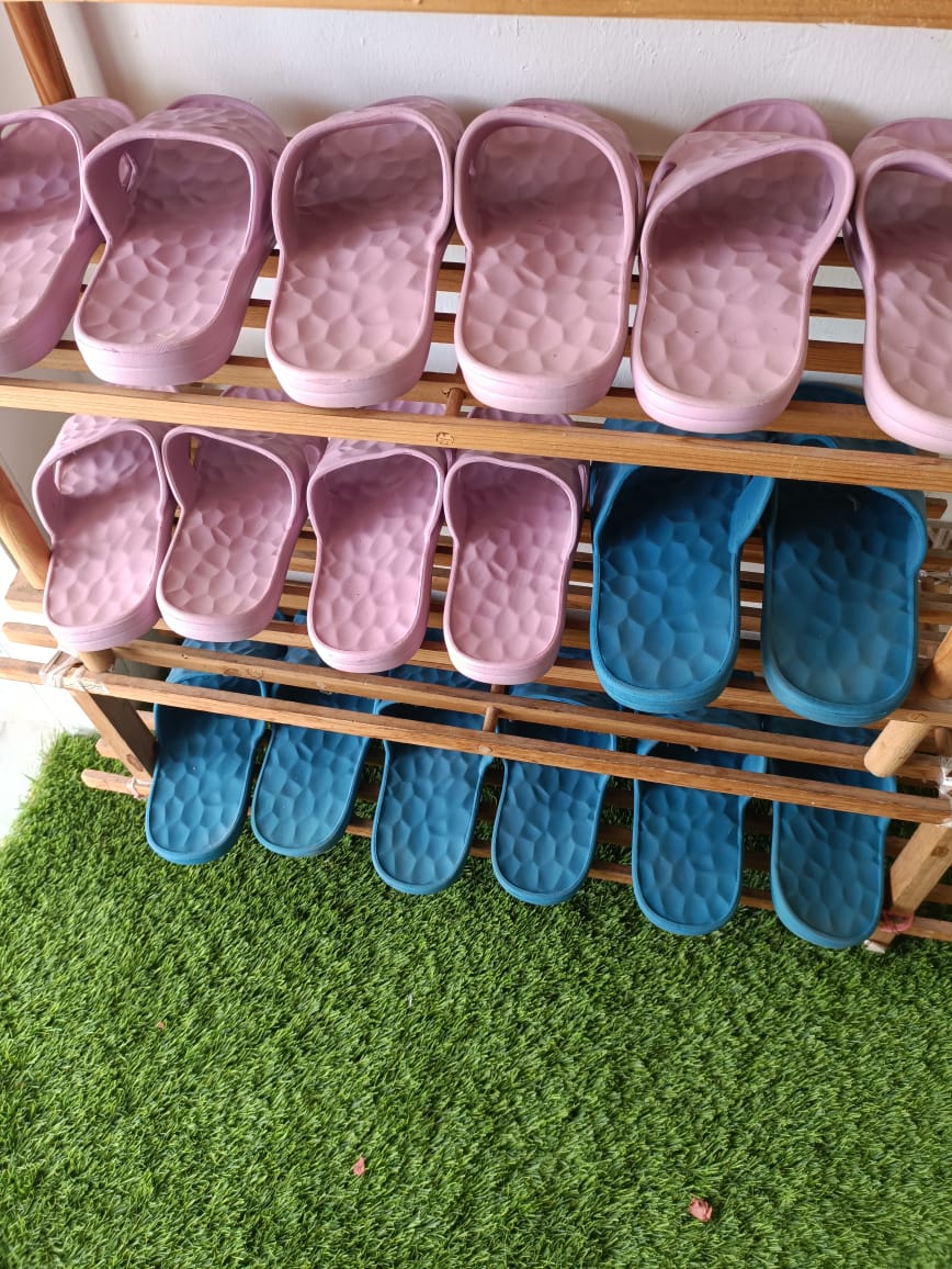 A neatly arranged shoe rack displays a variety of colorful slippers. The upper shelf features pink slippers, while the lower shelf holds blue slippers. The rack is positioned on a grassy surface, providing a welcoming touch to the area.