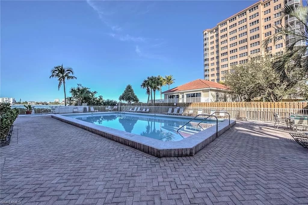 A landscaped pool area features a large swimming pool surrounded by a brick patio. Several lounge chairs are positioned around the pool, with palm trees and greenery providing a natural backdrop. A nearby building can be seen in the background.