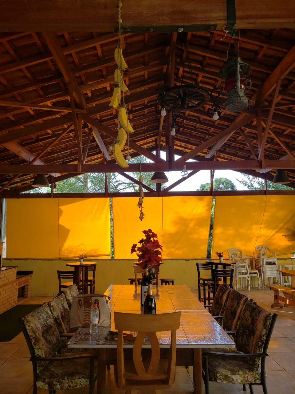 An outdoor dining area features a wooden ceiling supported by beams, with a large table at its center. Colorful flowers are placed as a centerpiece. Yellow shades filter natural light, while empty chairs are visible in the background. Ceiling decorations include hanging bananas and a lantern.