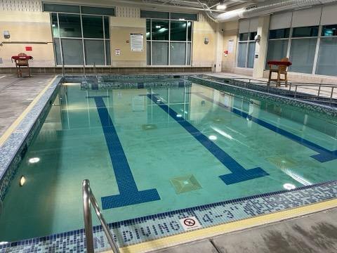 An indoor swimming pool features clear water with lane markings visible on the bottom. Surrounding windows allow natural light to fill the space, while guardrails provide safety access to the shallow end. Chairs are positioned nearby for convenience.
