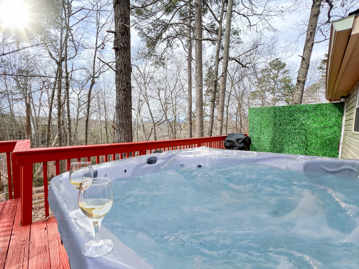 A hot tub sits on a red wooden deck, with two glasses positioned nearby. Soft sunlight filters through the trees, illuminating the serene forest views in the background, creating a peaceful atmosphere for relaxation.
