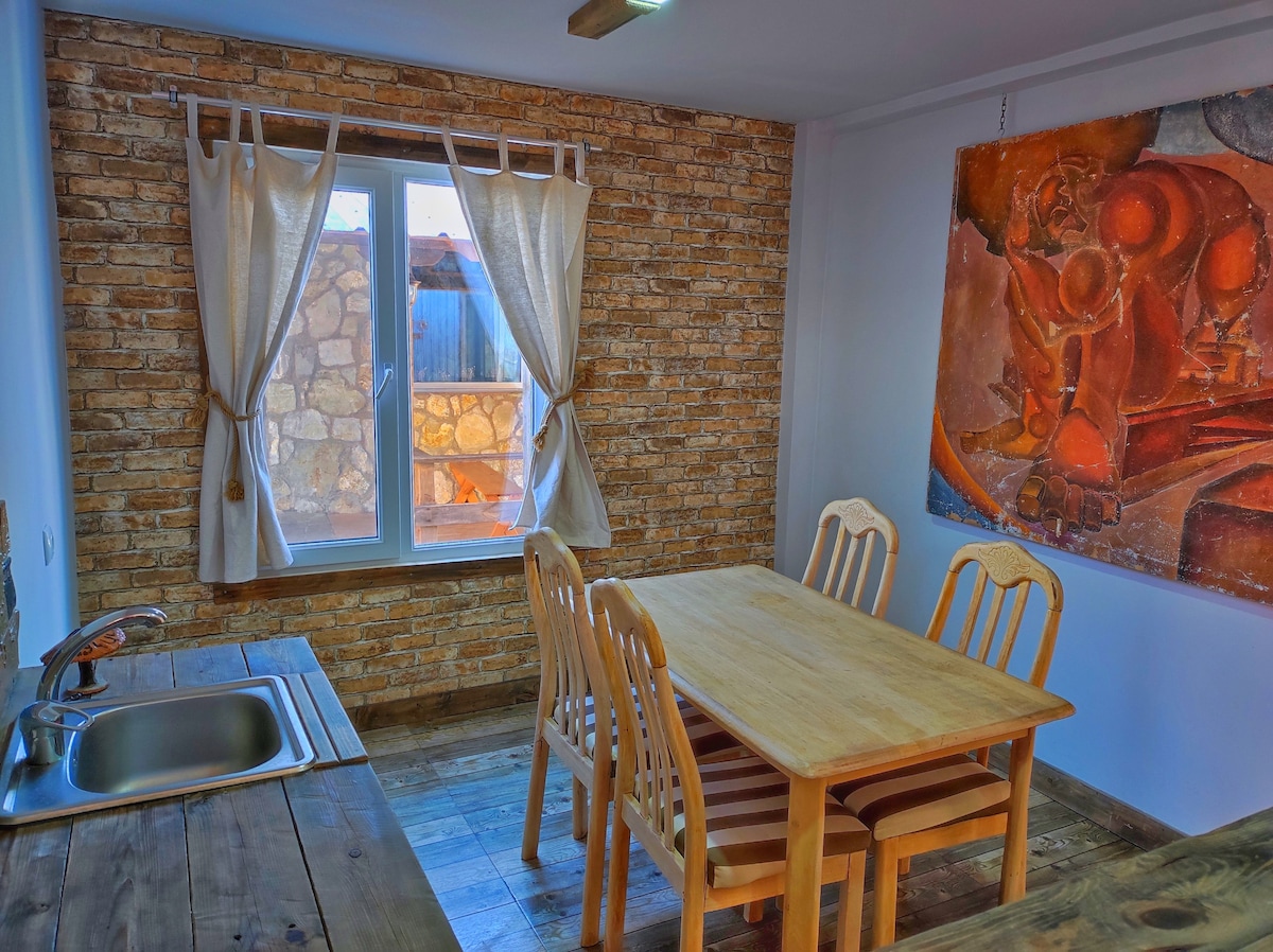 A dining area features a wooden table surrounded by six chairs. A window, dressed with light curtains, allows natural light to fill the space. A rustic stone wall adds texture, while an abstract artwork hangs nearby, providing a splash of color to the room.