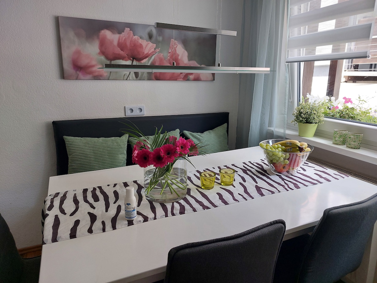 A dining table is adorned with a zebra-striped runner and features a vibrant arrangement of pink flowers. A bowl of fruit, including apples and grapes, is placed at the center. Soft seating is provided along one side, with a large window offering natural light.