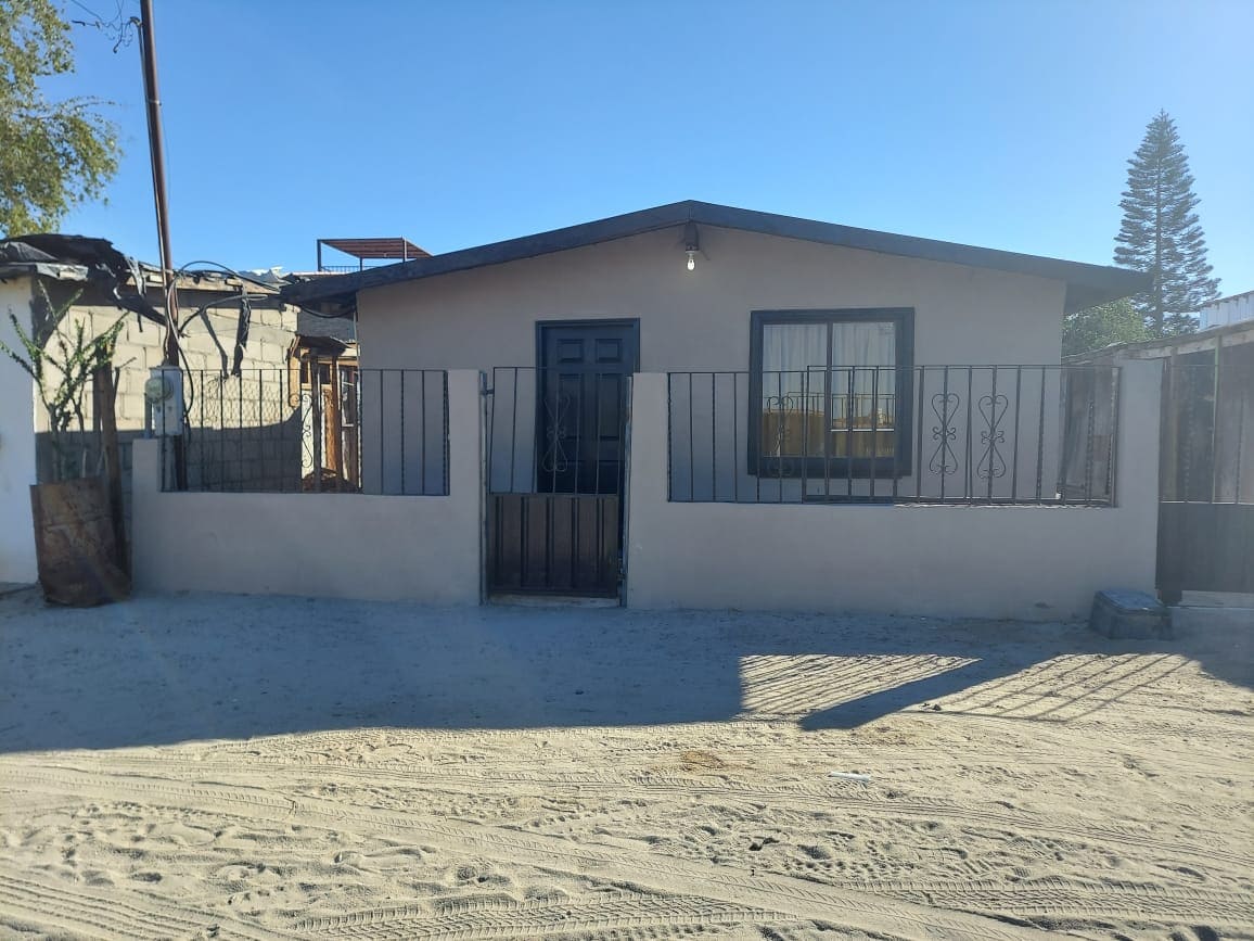 The front of a modest house is visible, featuring a dark door framed by a large window. A simple security fence surrounds the property, with sand underfoot and some greenery in the background. The clear blue sky provides a bright backdrop.